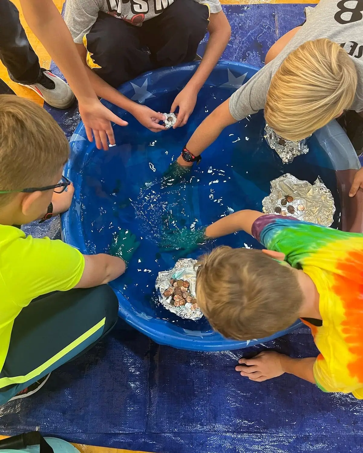 Children arrange foil boats and coins in a blue tub of water during a hands-on activity.