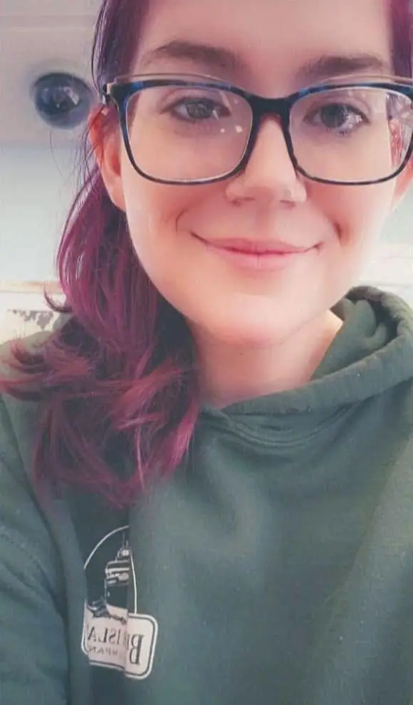 Close-up portrait of a smiling woman with pink-purple hair, black glasses, and a dark green hoodie.