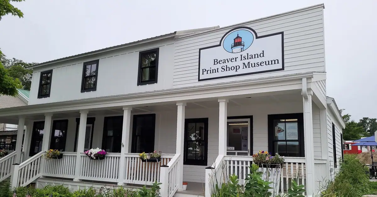 White wood-sided building with a porch, flower boxes, and a large museum sign bearing a lighthouse logo.