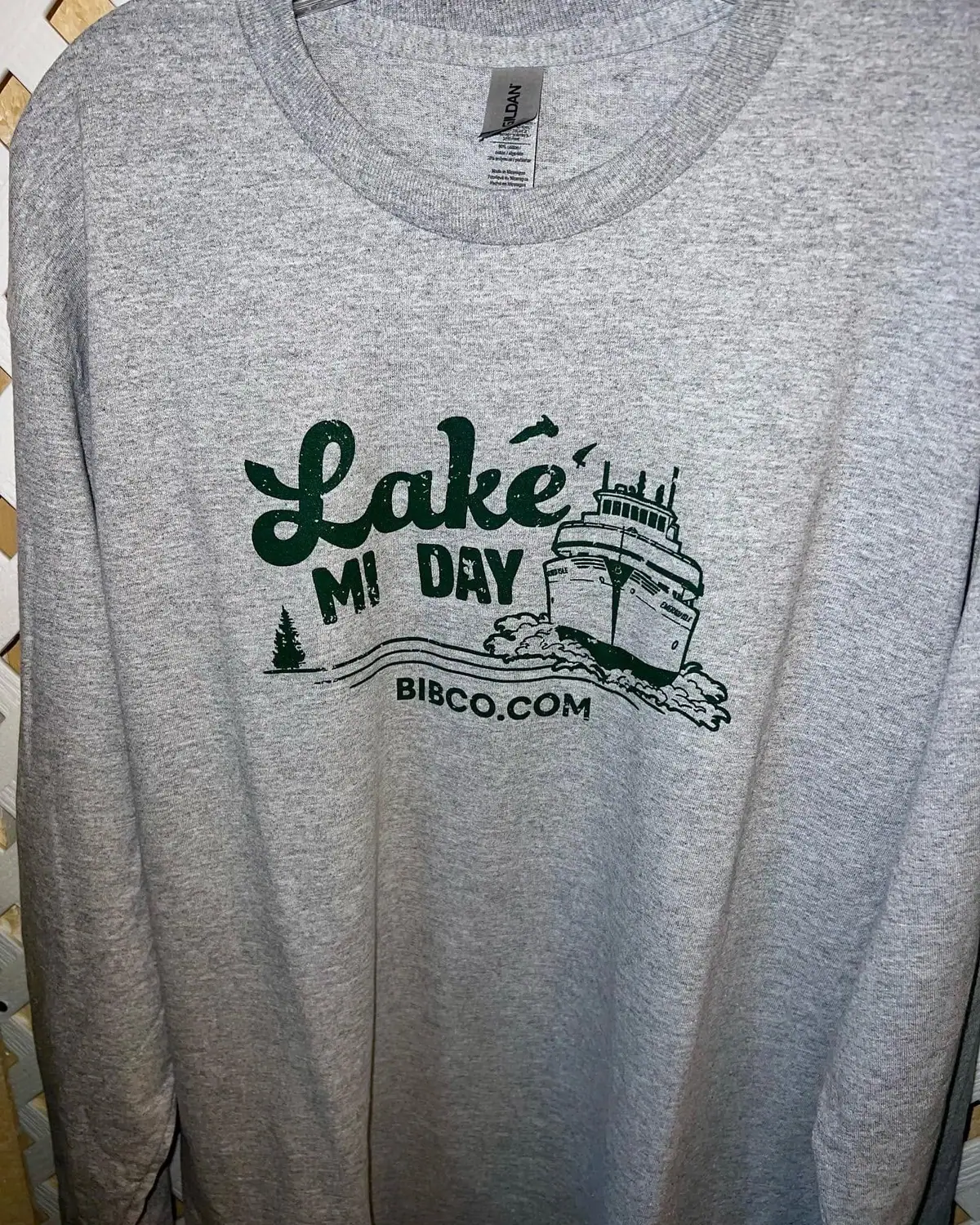 Gray long-sleeved shirt displaying a dark green graphic of a passenger vessel, waves, an evergreen tree, and lettering.
