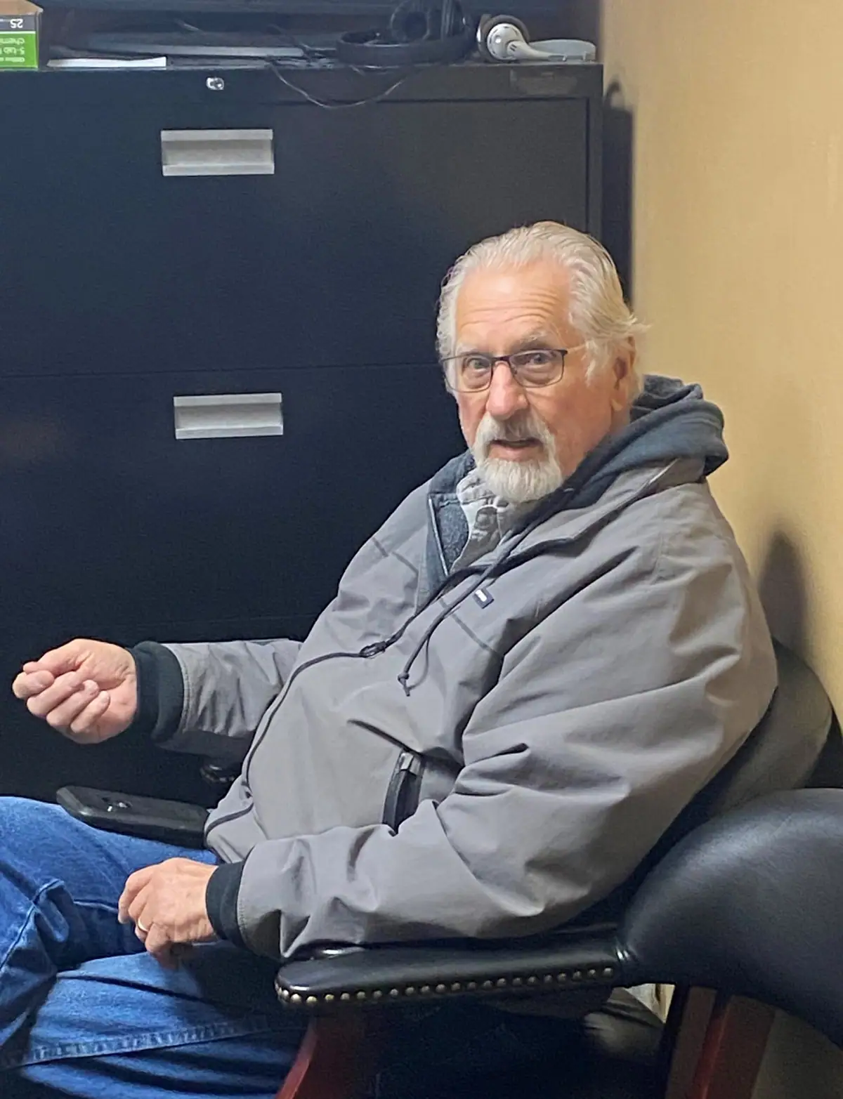 A bearded man in glasses sits in an office chair beside a dark filing cabinet.