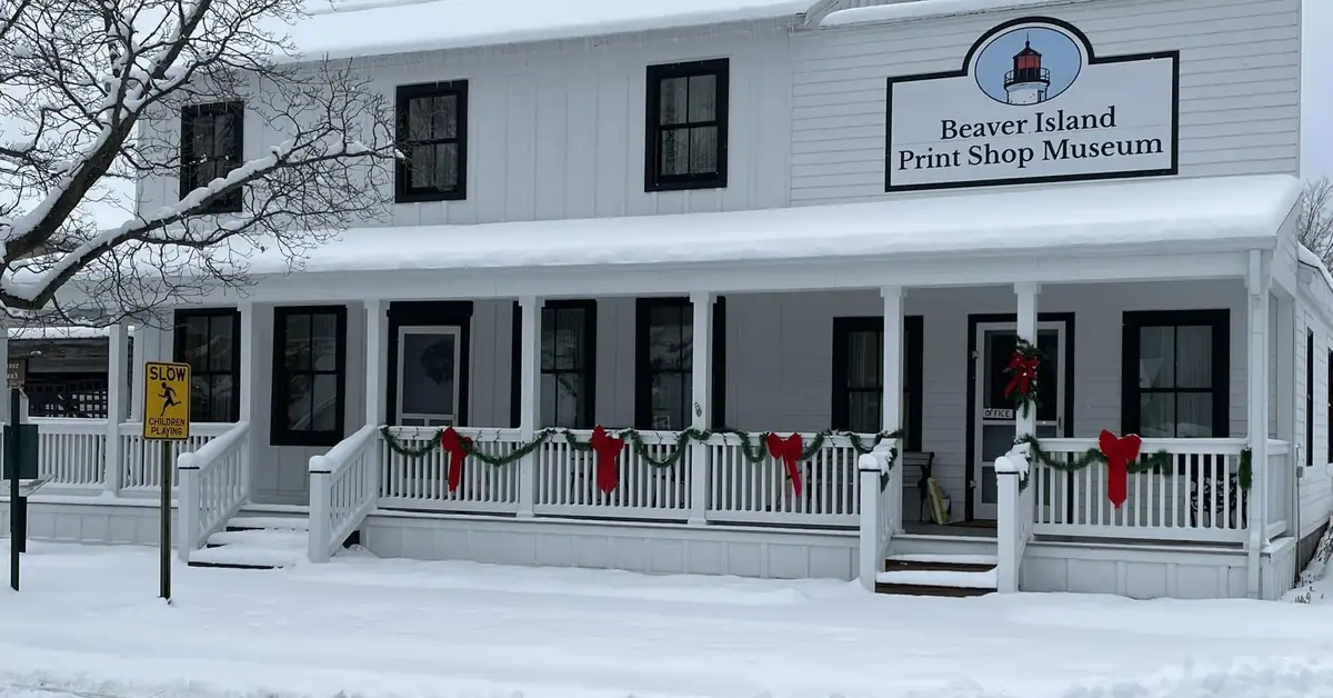 Snow-covered white building with a porch, holiday garlands, and a sign for the Beaver Island Print Shop Museum.