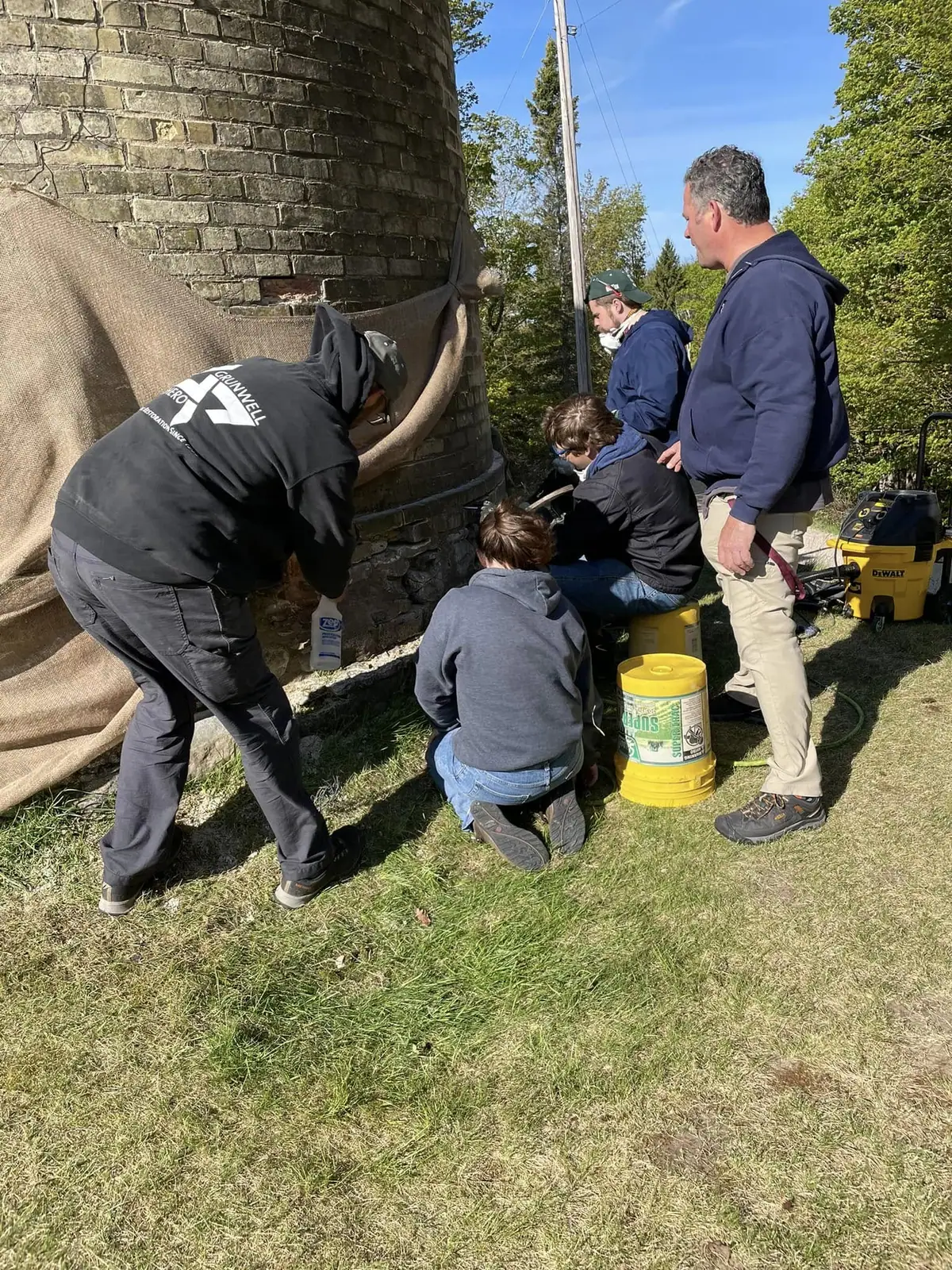 Five people repair a cylindrical brick chimney outdoors beside masonry supplies and power tools.