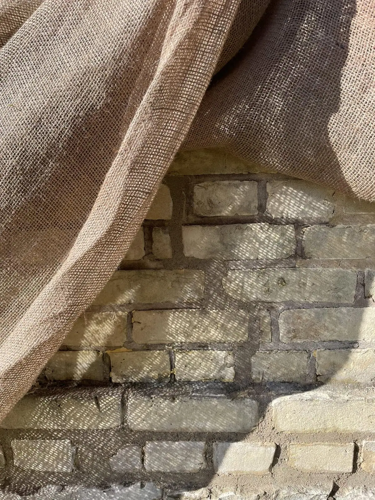 A coarse woven cloth drapes over a sunlit brick wall.