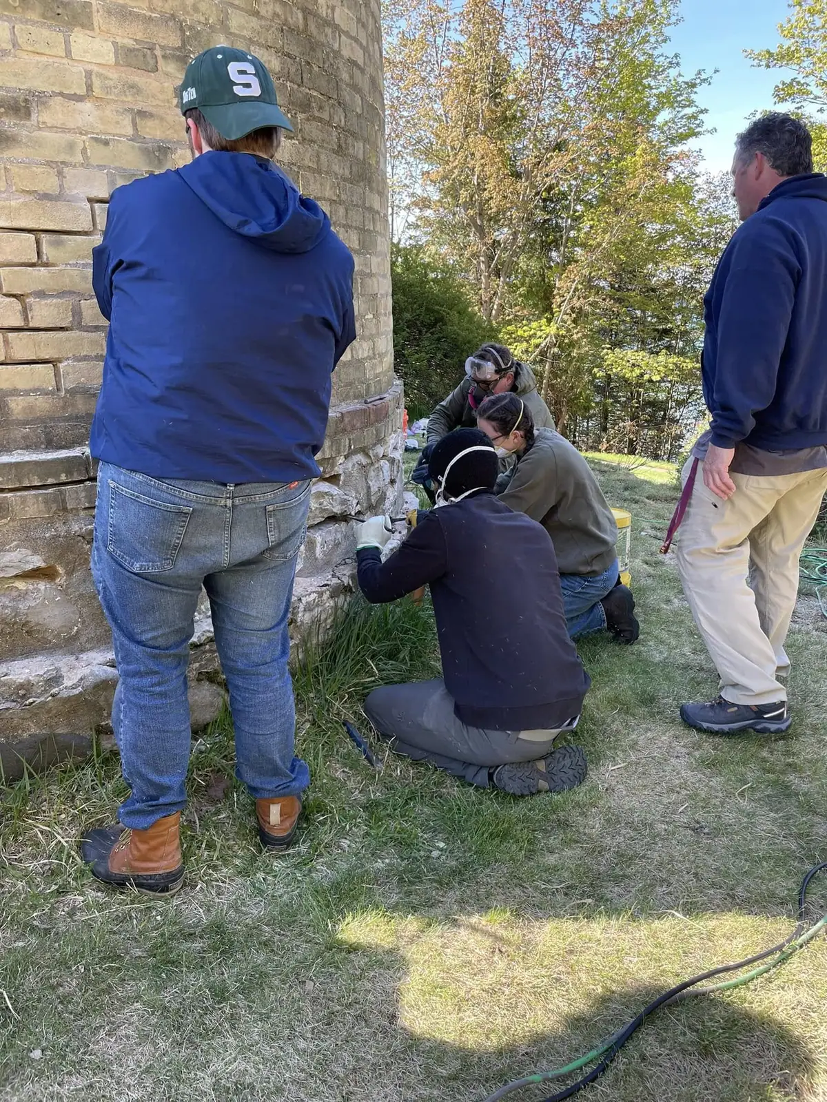 Five people work with tools at the base of a cylindrical brick tower outdoors.