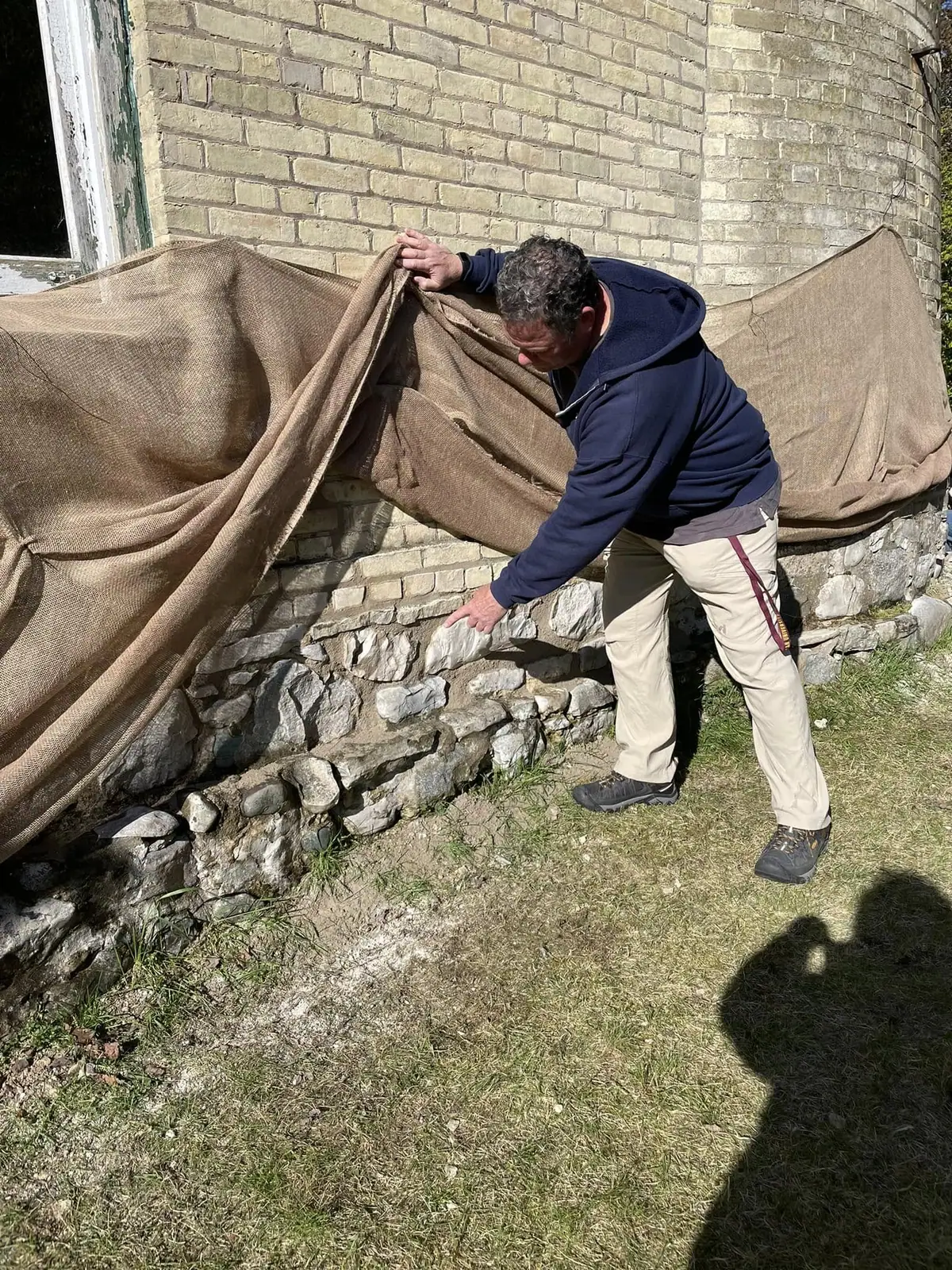 A man lifts a burlap tarp to inspect the stone base of a brick wall.