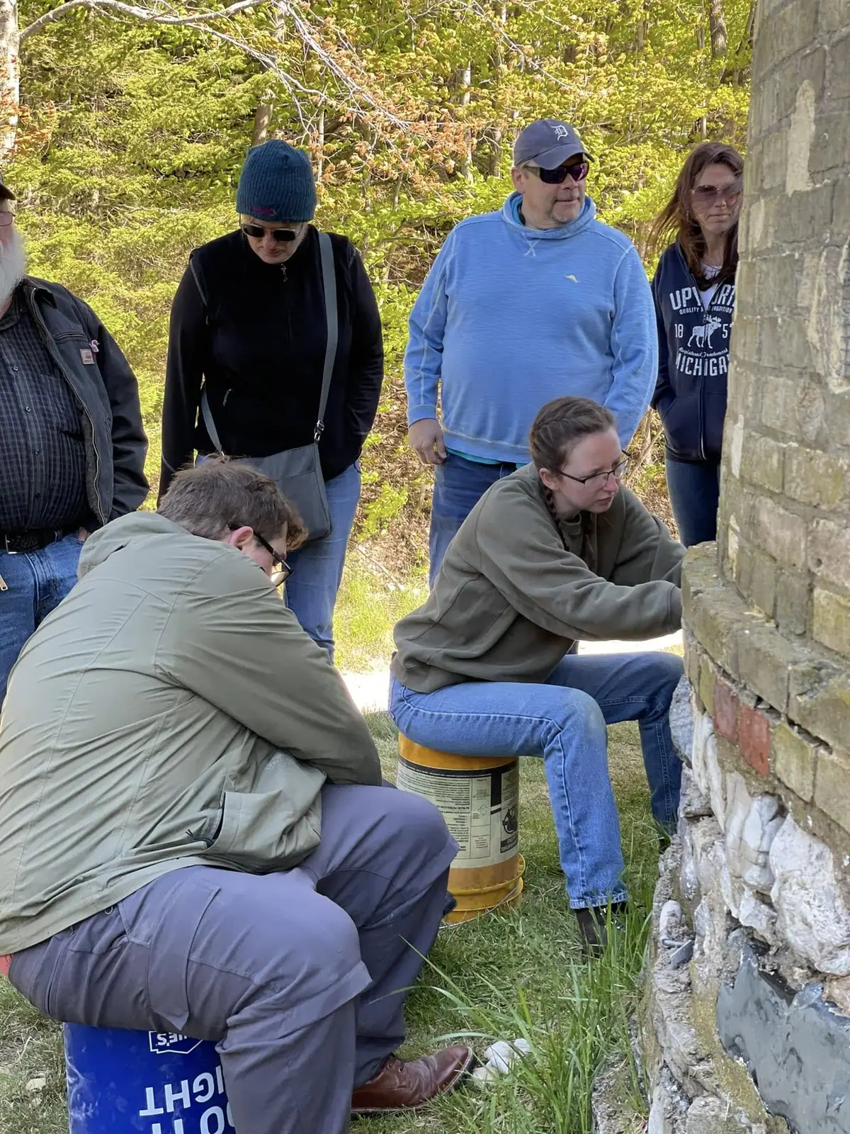 Six people gather beside a stone-and-brick wall in a wooded setting, with two seated on plastic barrels.