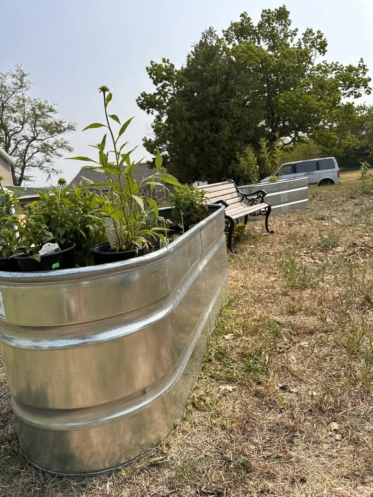 Galvanized raised planters filled with potted plants beside a bench, trees, houses, and a parked van.