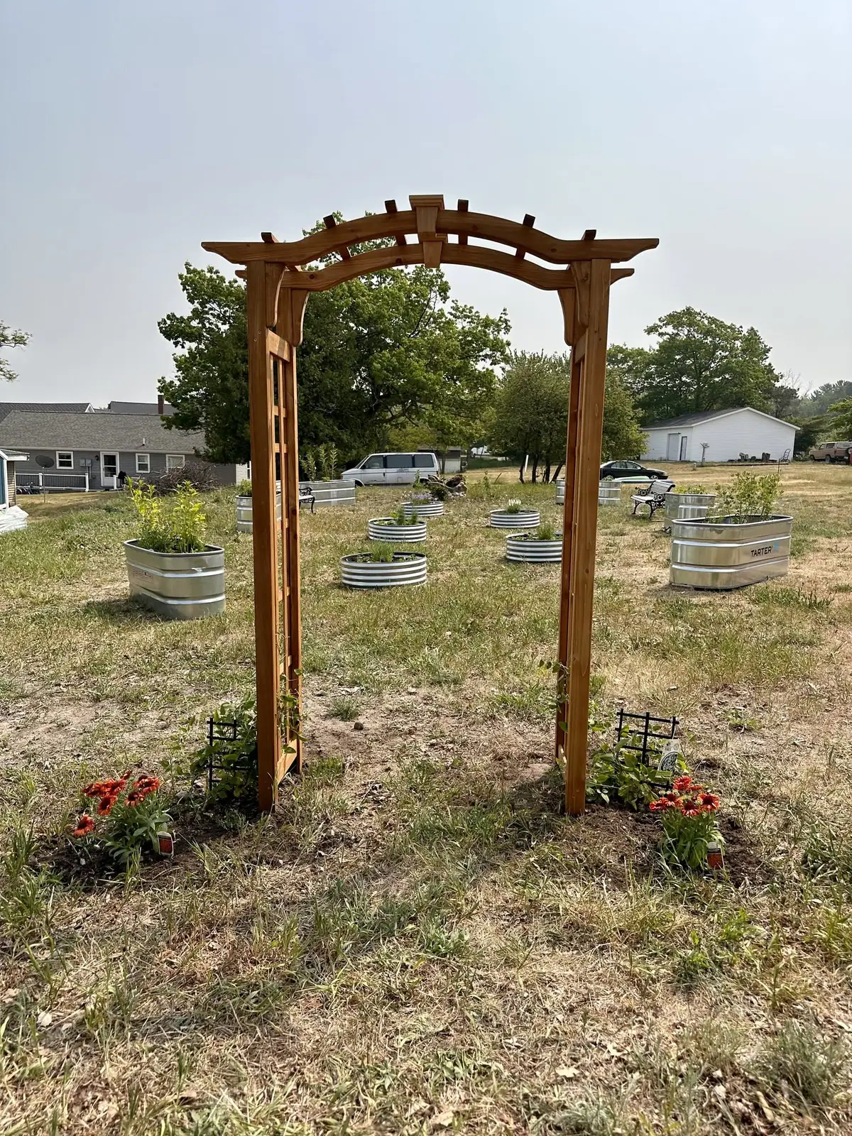 A wooden garden arbor stands among metal planters and flowers in a grassy yard.