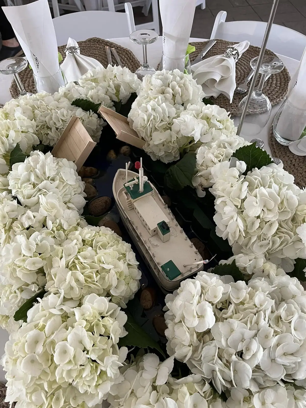A table centerpiece of white hydrangeas surrounds a model vessel and small wooden boats.