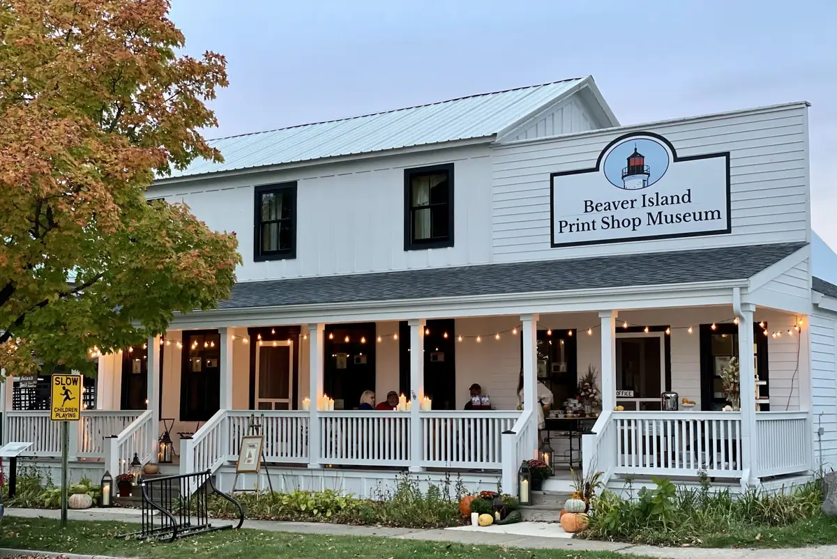 White clapboard building with a porch, illuminated sign, string lights, and autumn decorations.