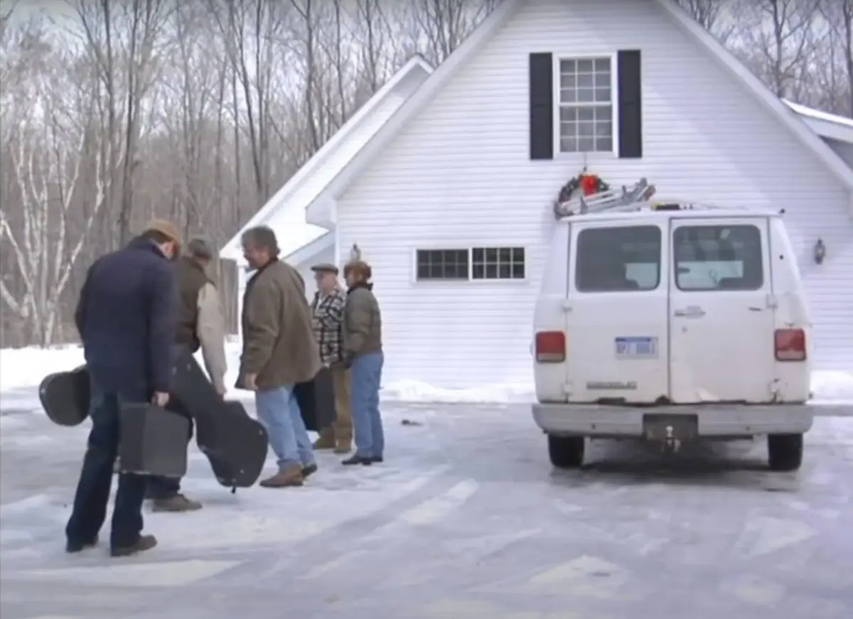 People carrying instrument cases stand beside a white van and house in a snowy driveway.