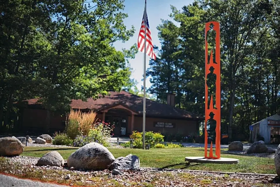 A low brown building stands among trees, with a flagpole and a tall orange sculpture of three figures.