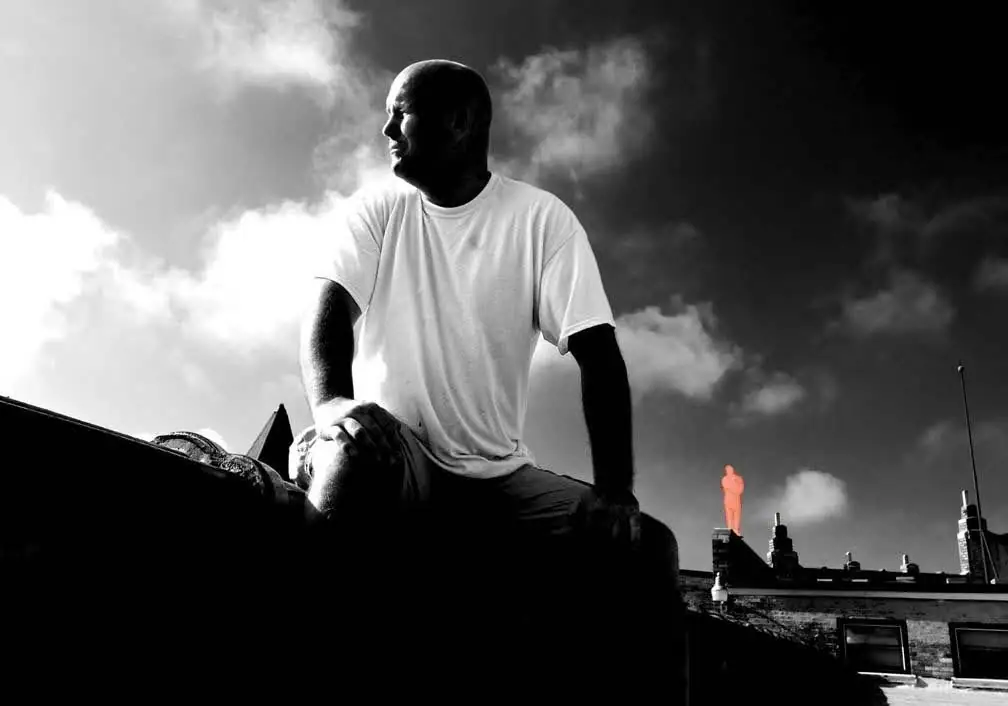A man in a white shirt sits on a rooftop ledge beneath dramatic clouds.