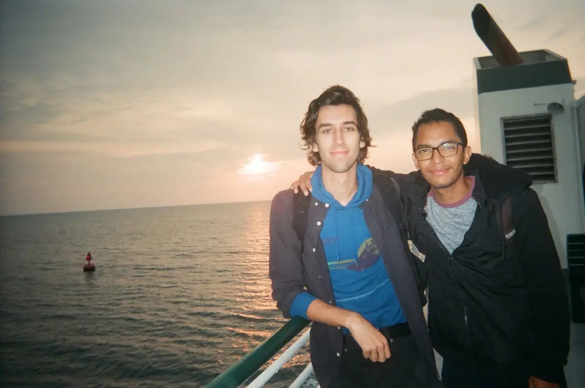 Two young men pose on a vessel’s deck with open water, a buoy, and the setting sun behind them.