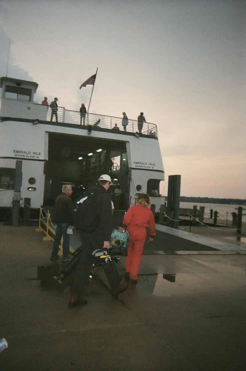 A white ferry is open at a wet pier as passengers walk aboard in the fading light.