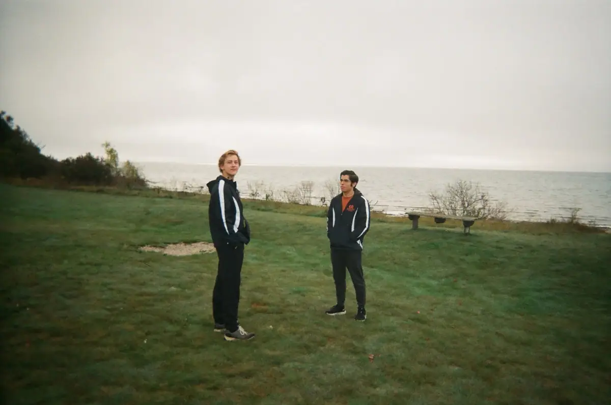 Two young men stand on a grassy waterfront beside a wooden bench beneath an overcast sky.