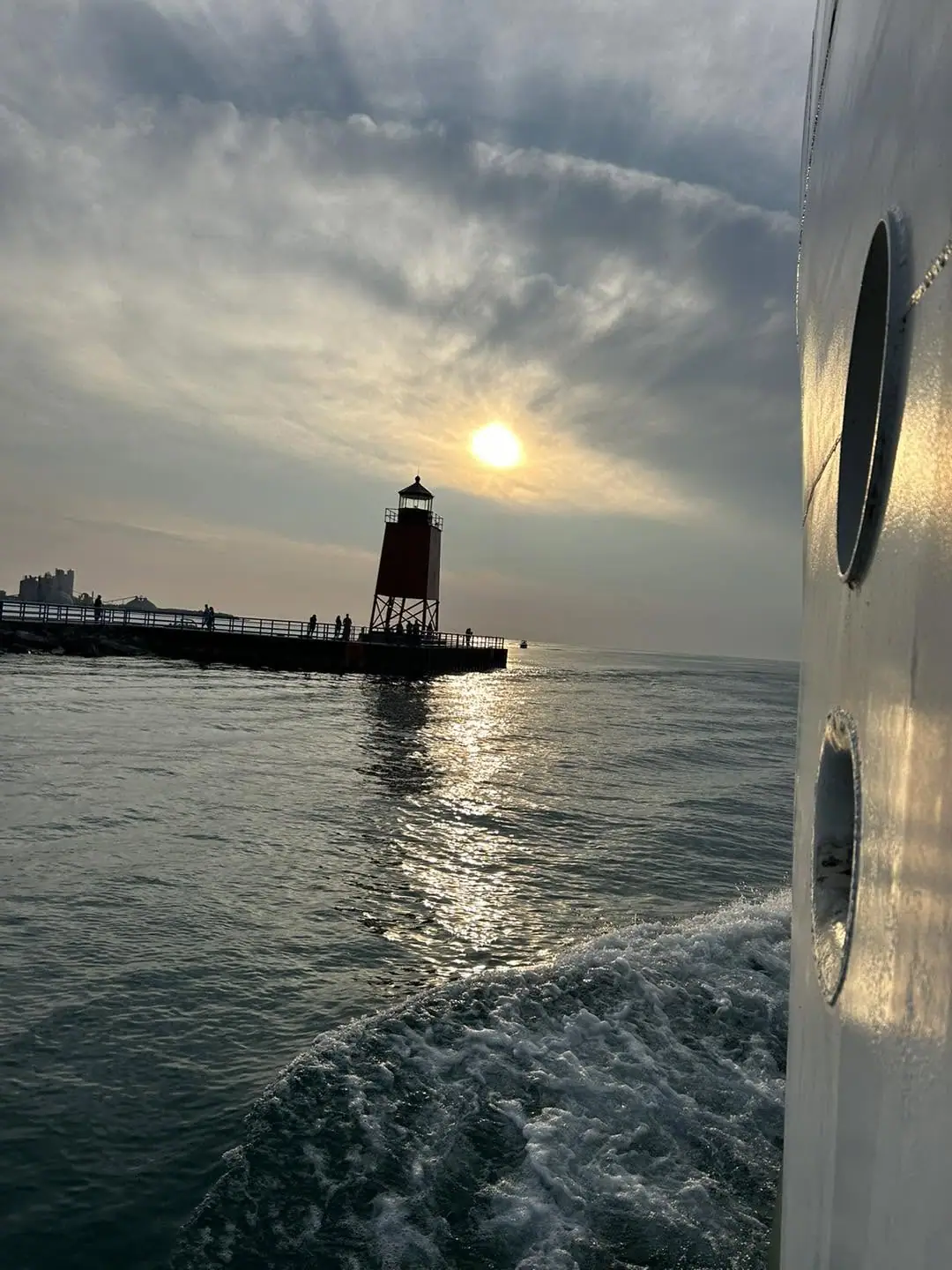 A lighthouse stands at the end of a pier as a boat passes through sunlit water at sunset.