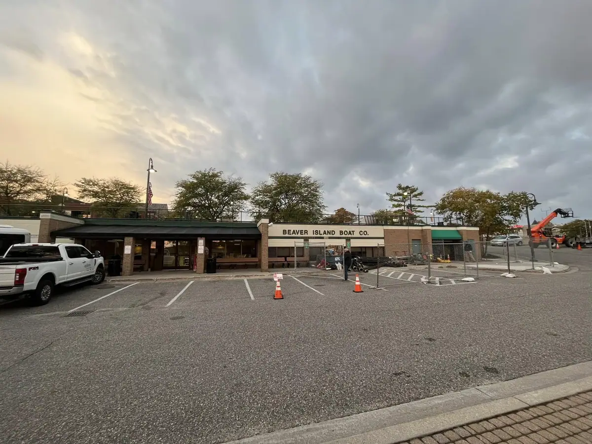 A boat company terminal with a sign, parked vehicles, cones, and an orange lift truck beneath a cloudy sky.