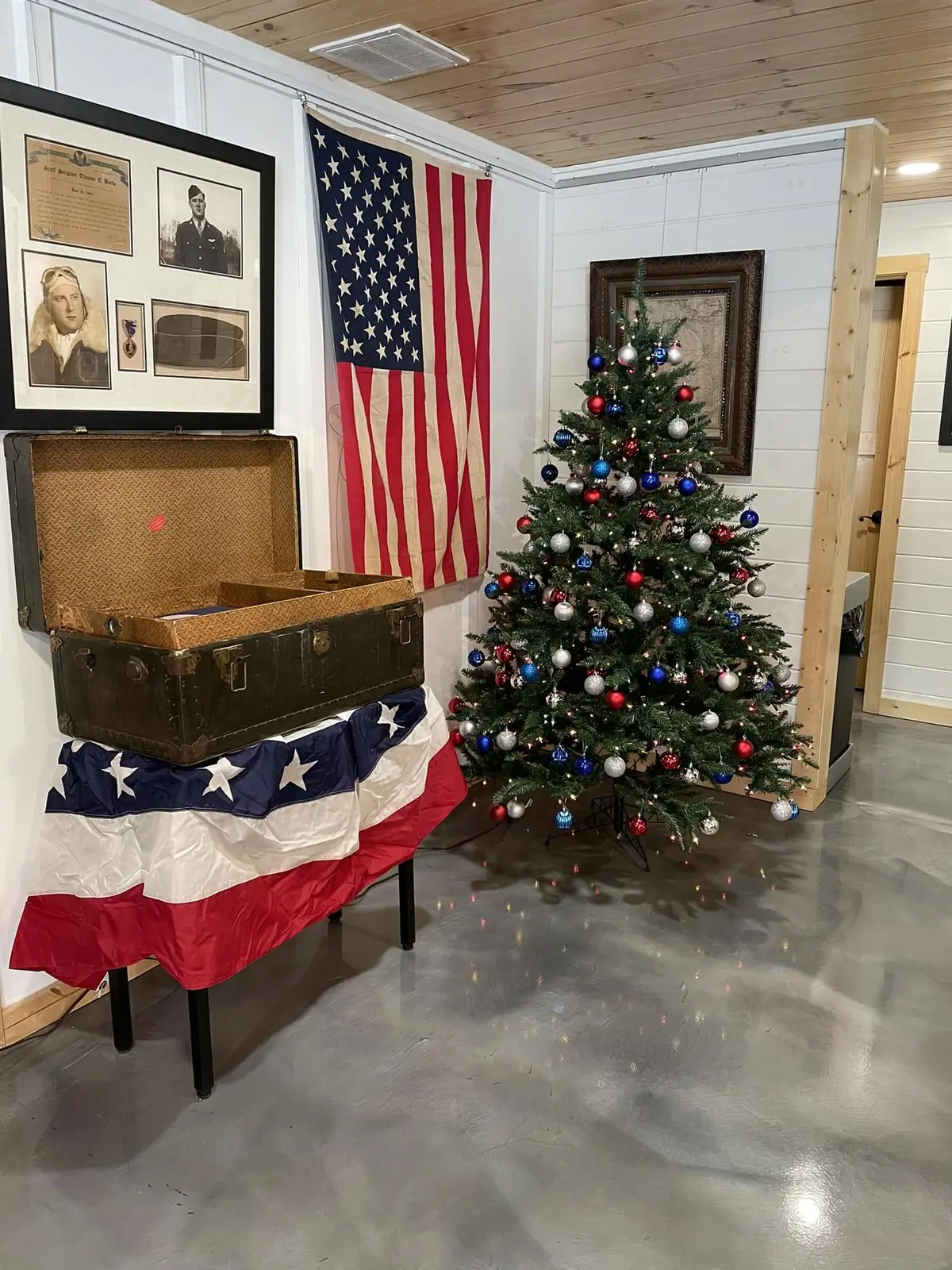 Indoor museum display with a military portrait, American flag, open suitcase, and decorated Christmas tree.
