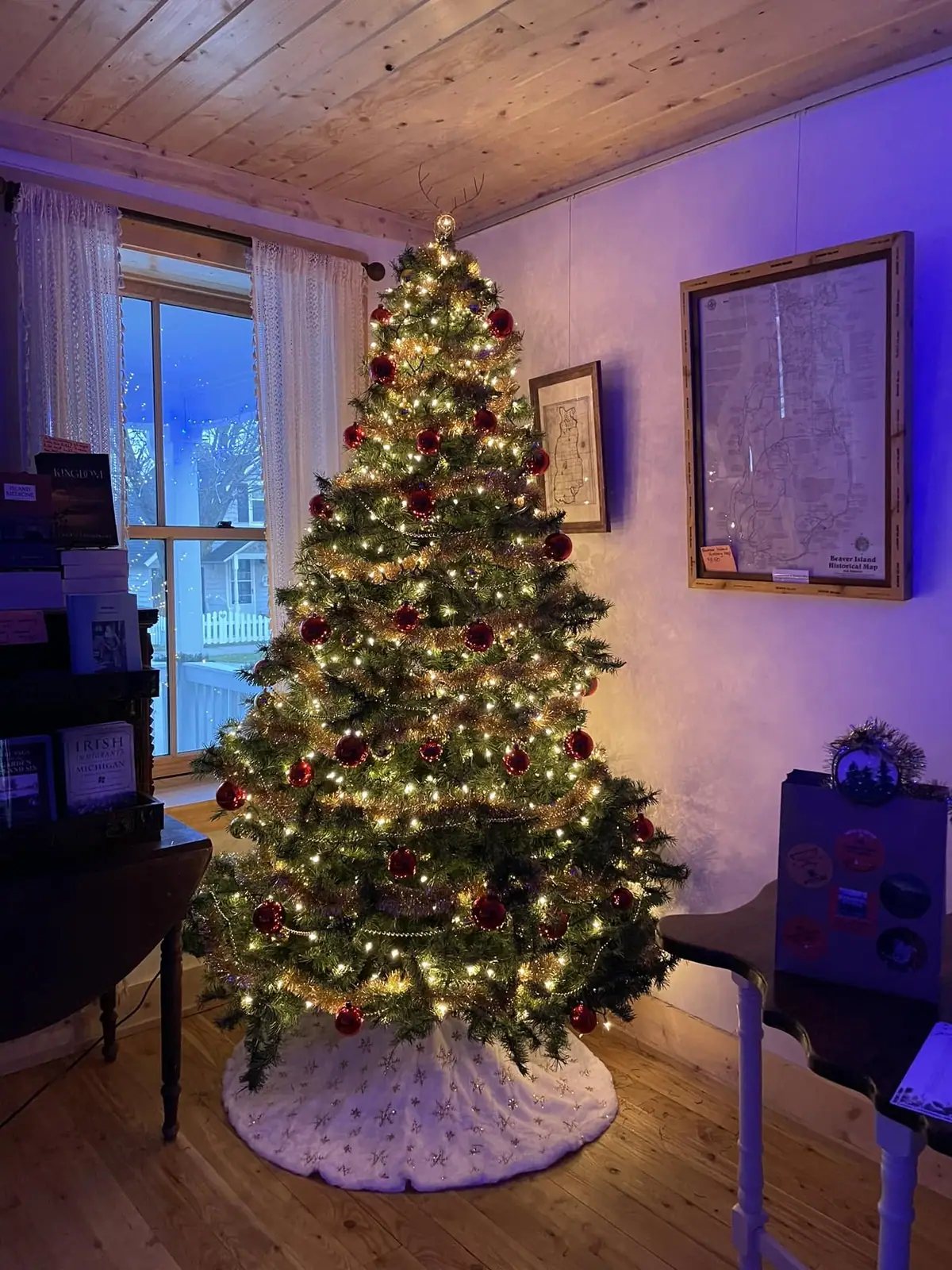 A lit Christmas tree decorated with red ornaments stands in a wood-paneled room beside framed maps.