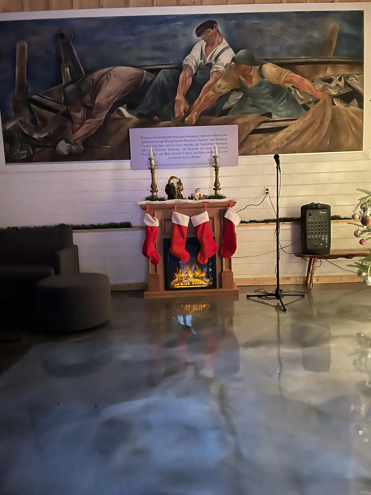 Interior decorated for the holidays, with a mural of workers above a fireplace and stockings.