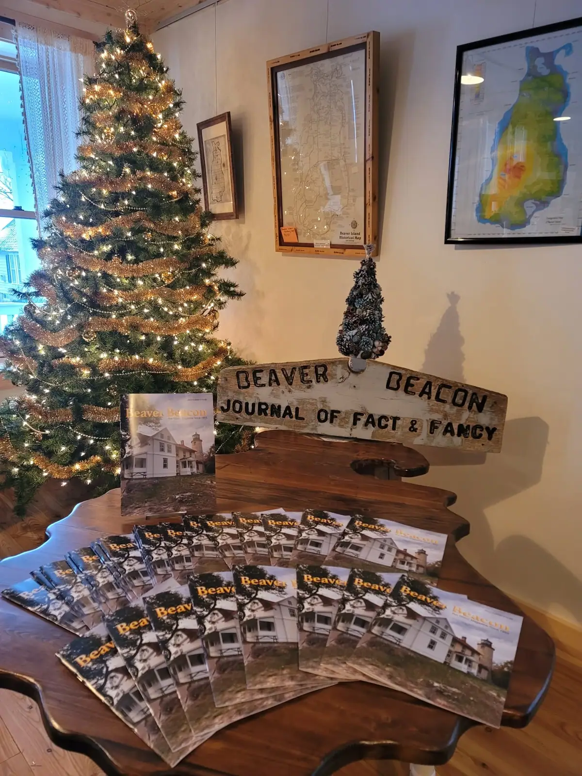 A decorated Christmas tree and magazines displayed on a wooden table beneath framed maps and a wooden sign.