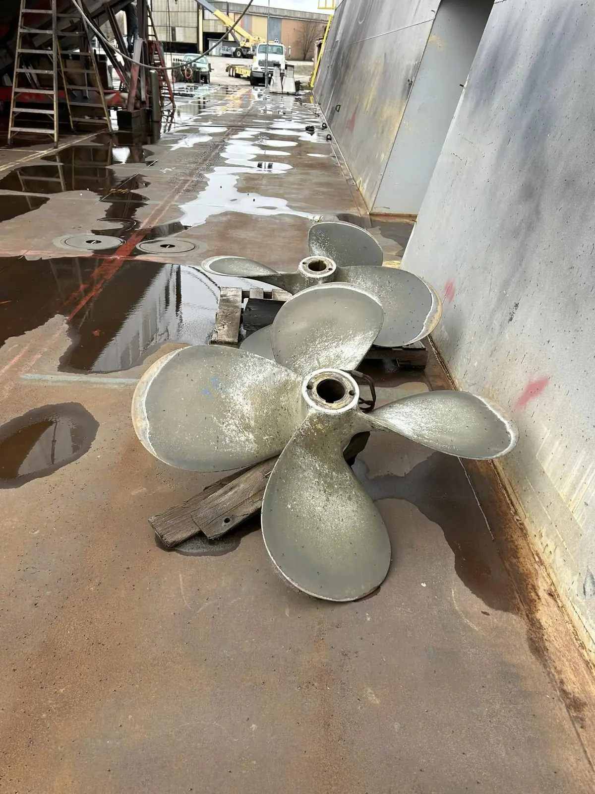 Two large three-bladed propellers lie on a wet industrial deck beside a vessel hull.