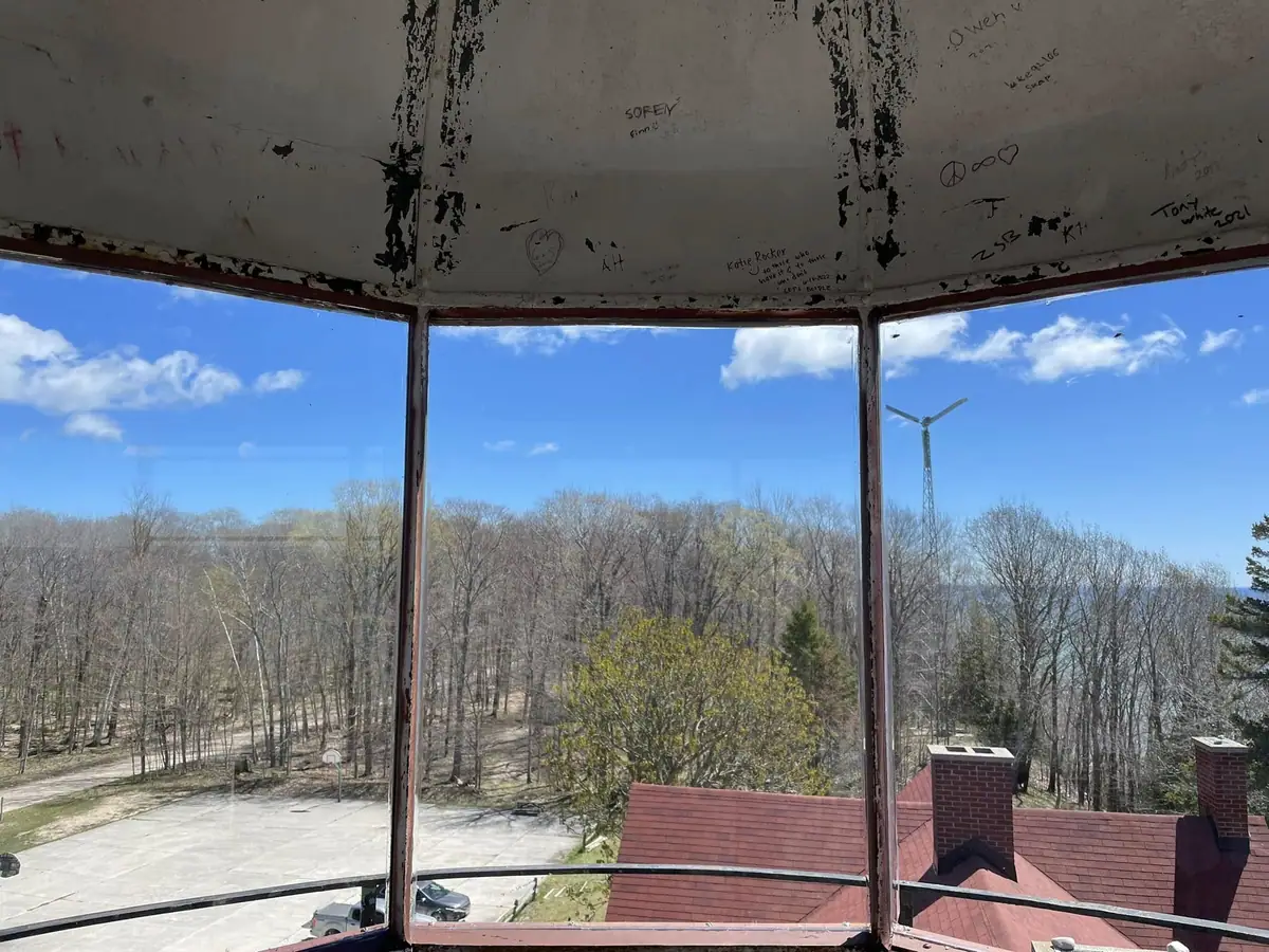 Graffiti-covered interior windows overlook leafless woods, a paved drive, rooftops, and a wind turbine.