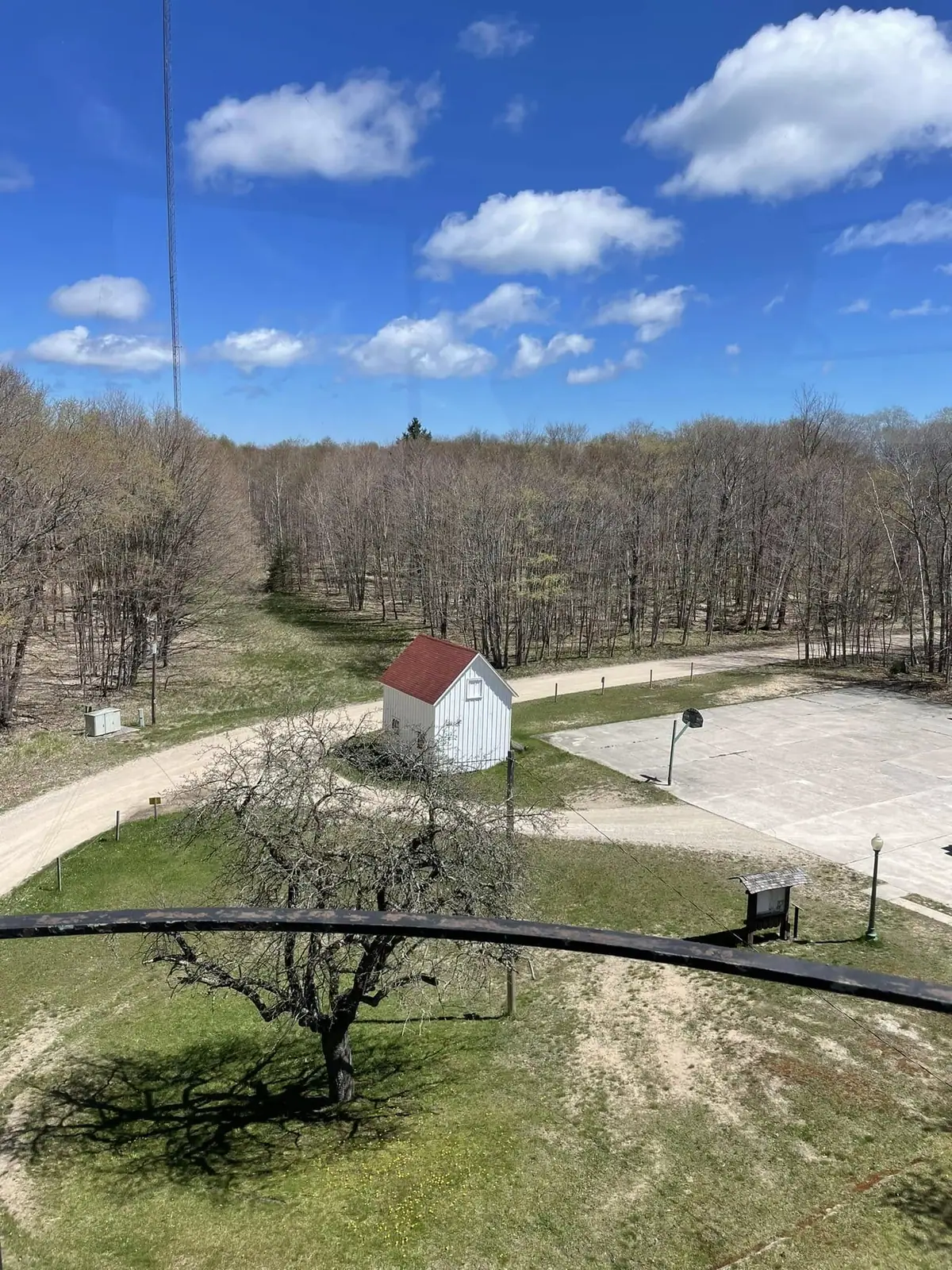 Elevated view of a white shed, basketball court, wooded grounds, and antenna tower under a blue sky.