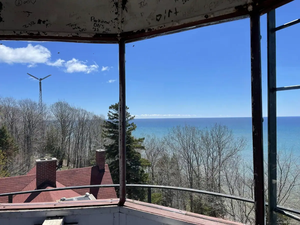 View through a weathered observation structure toward blue water, a forest, a red-roofed building, and a wind turbine.