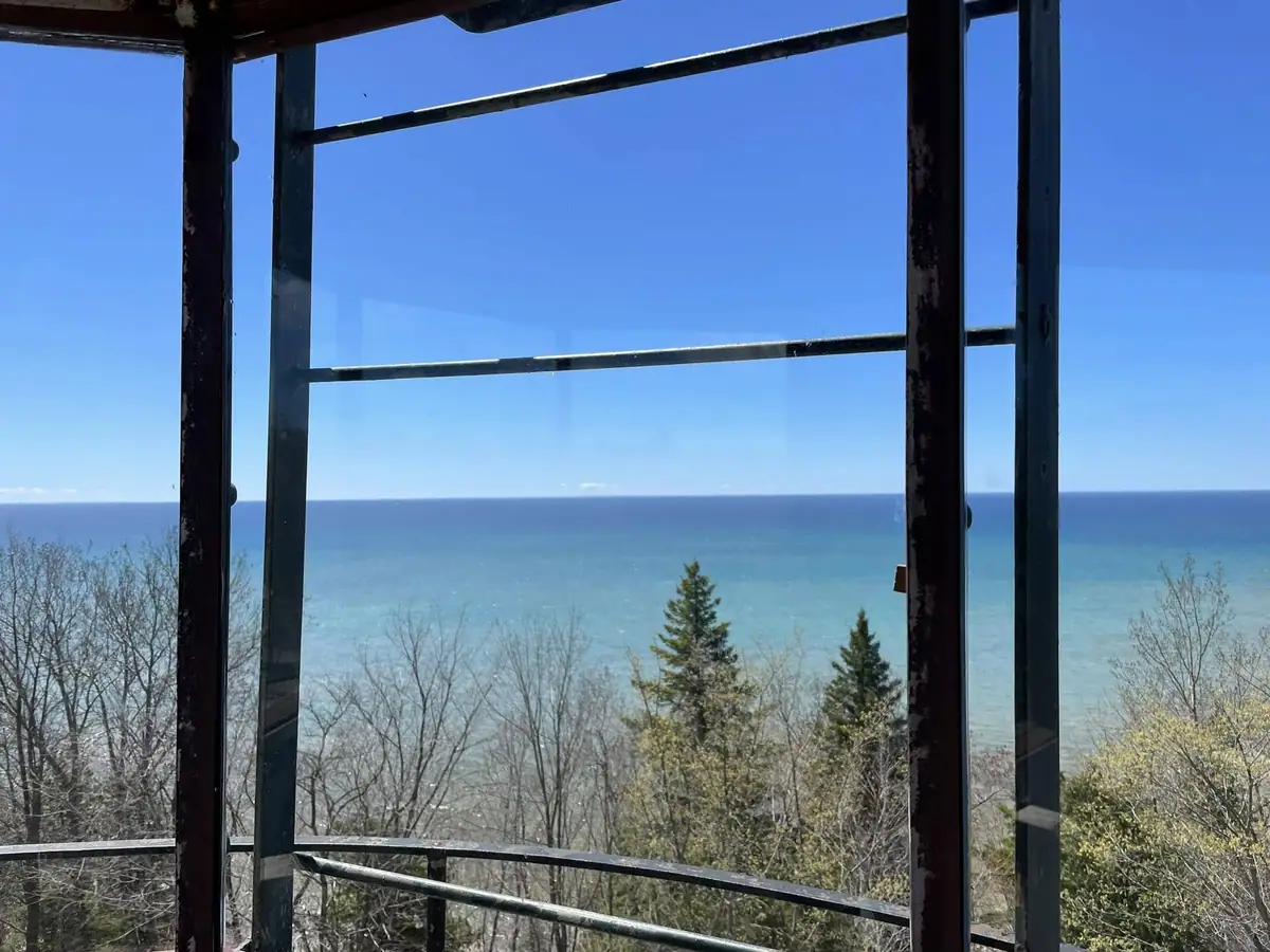 A broad blue body of water viewed through a weathered dark metal framework above leafless and evergreen trees.
