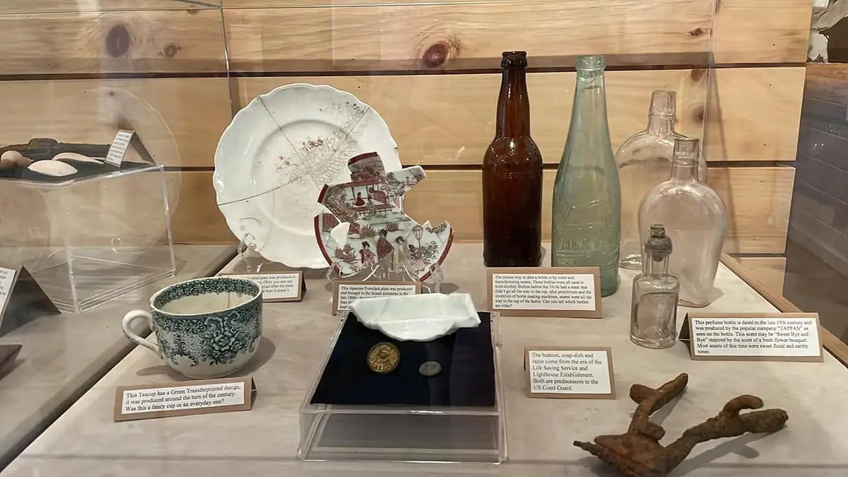 Museum display case holding decorated dishes, bottles, buttons, a soap dish, and a rusted anchor.
