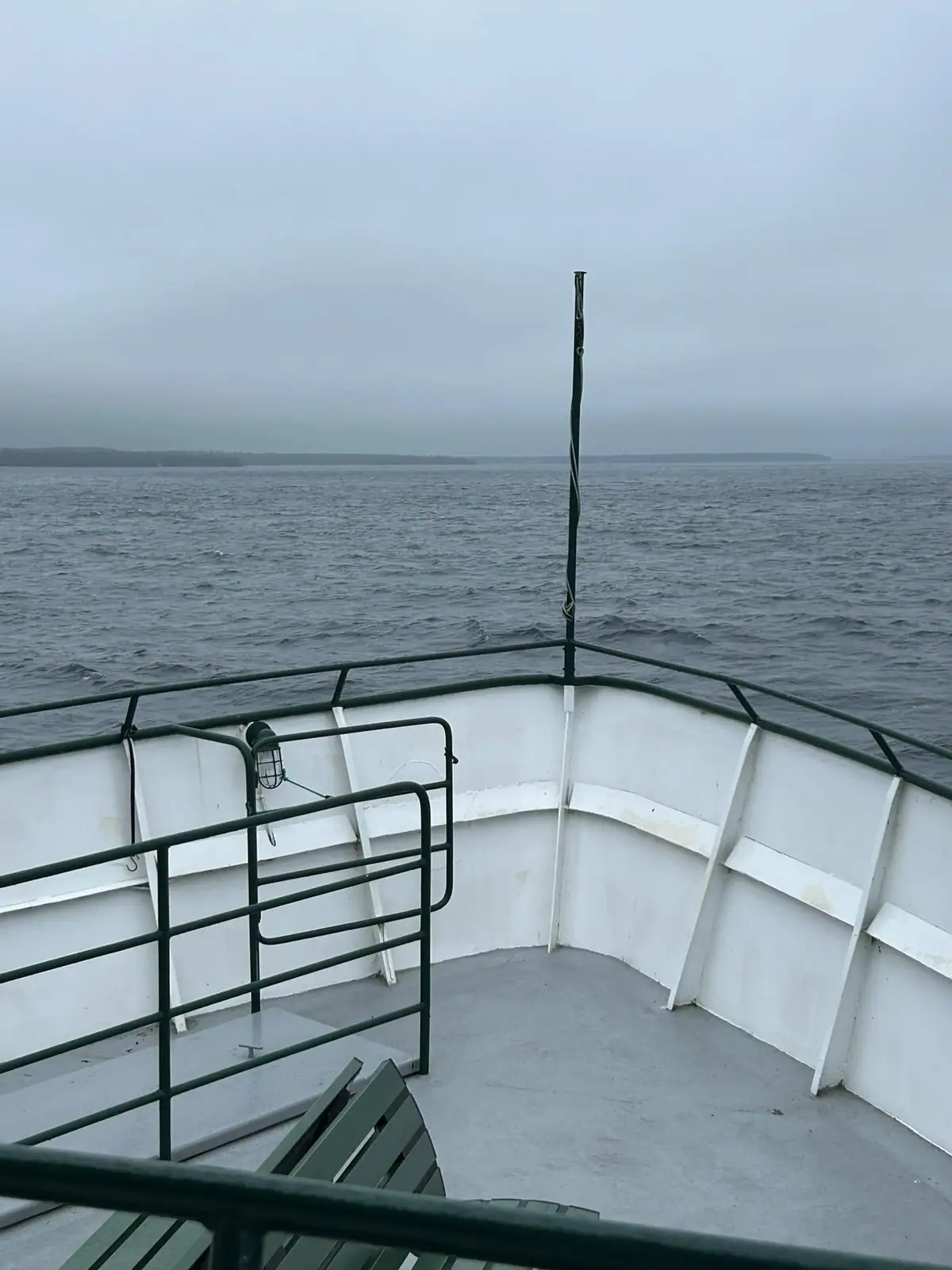 View over a ferry’s bow toward a low shoreline beneath an overcast sky.