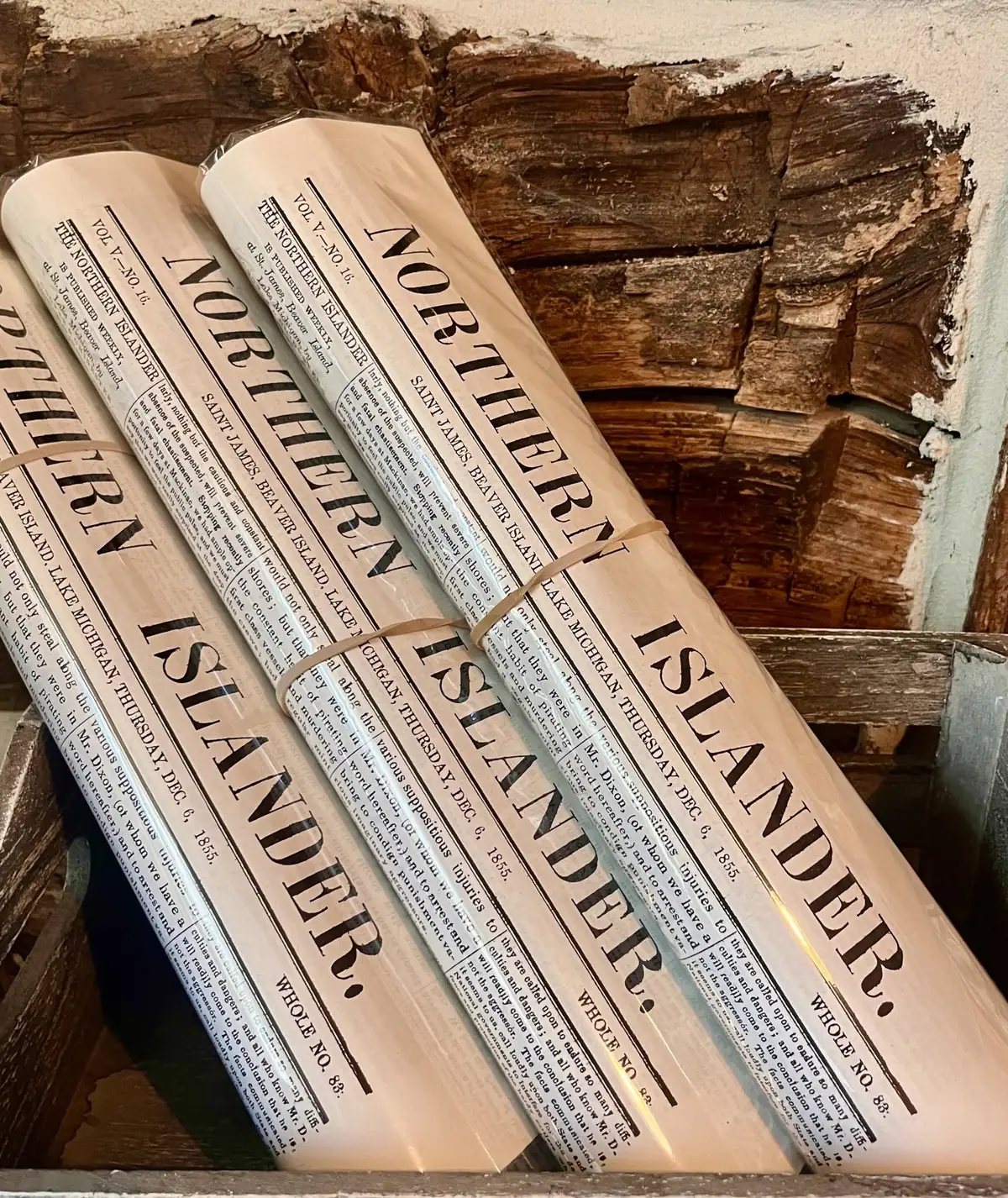 Several rolled newspapers rest against an exposed timber wall, with large mastheads and dense printed columns visible.