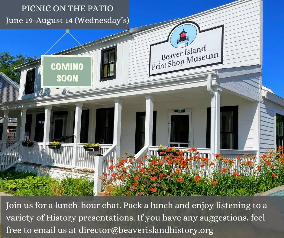 A white wood-sided building with a covered porch, museum sign, event lettering, and colorful flowers.