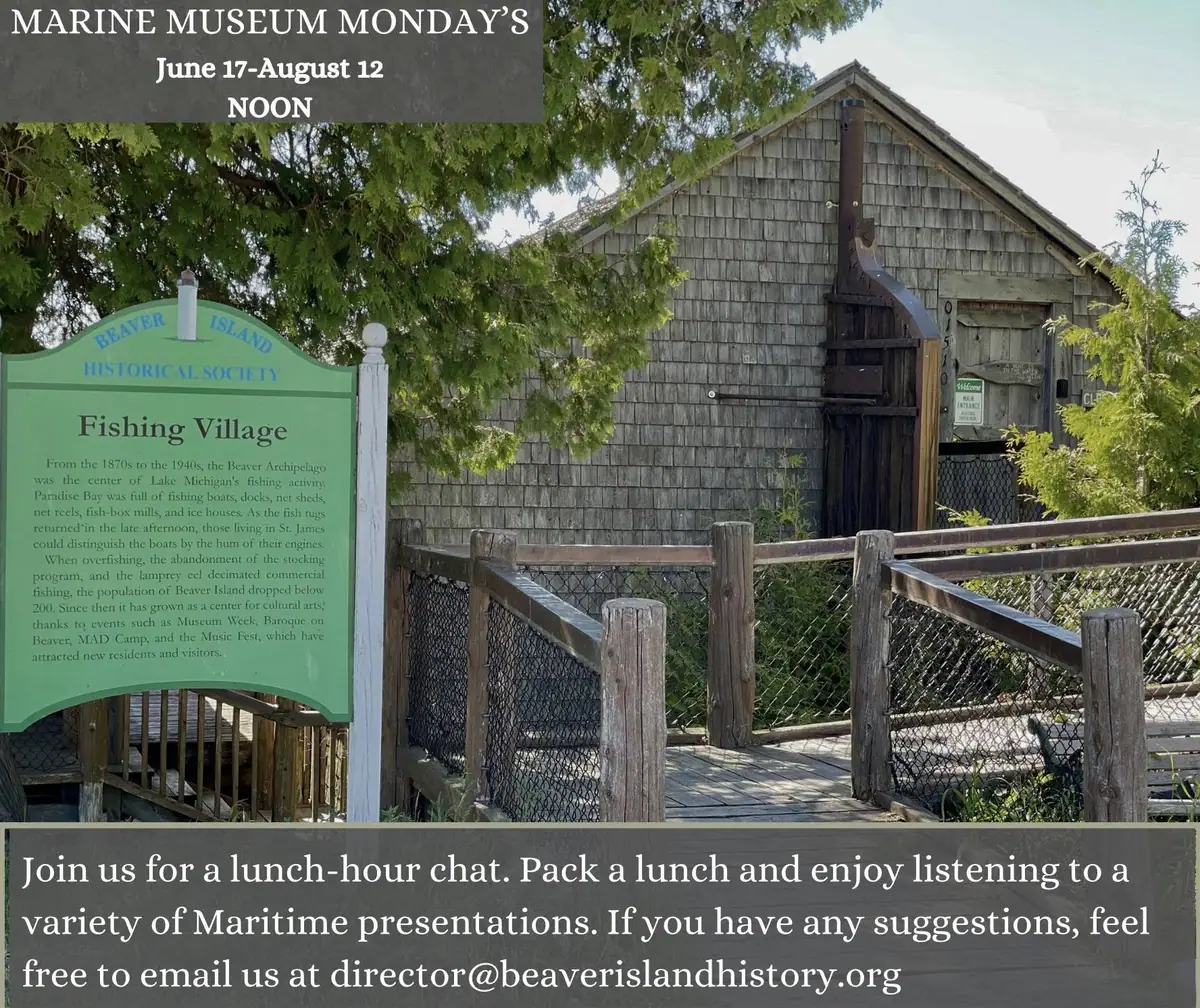 A green historical marker stands beside a shingled wooden building and fenced boardwalk.
