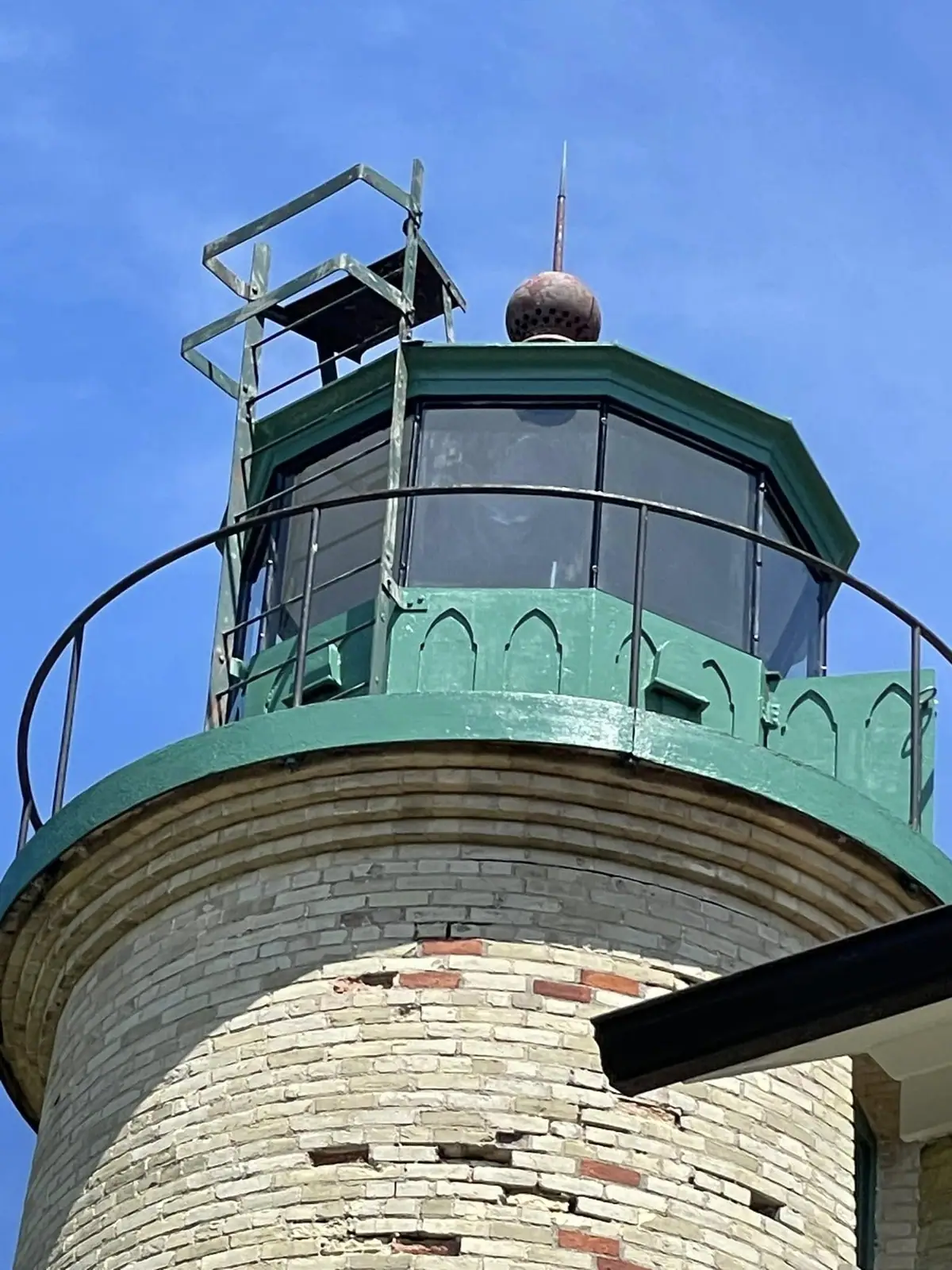 Close-up view of a weathered brick lighthouse tower and green lantern room against a blue sky.