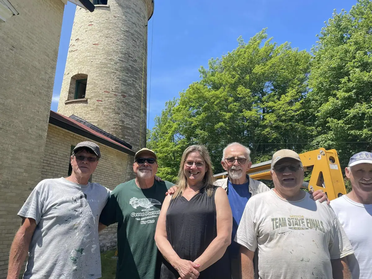 Six adults pose beside a pale brick lighthouse tower, with a yellow excavator and trees behind them.