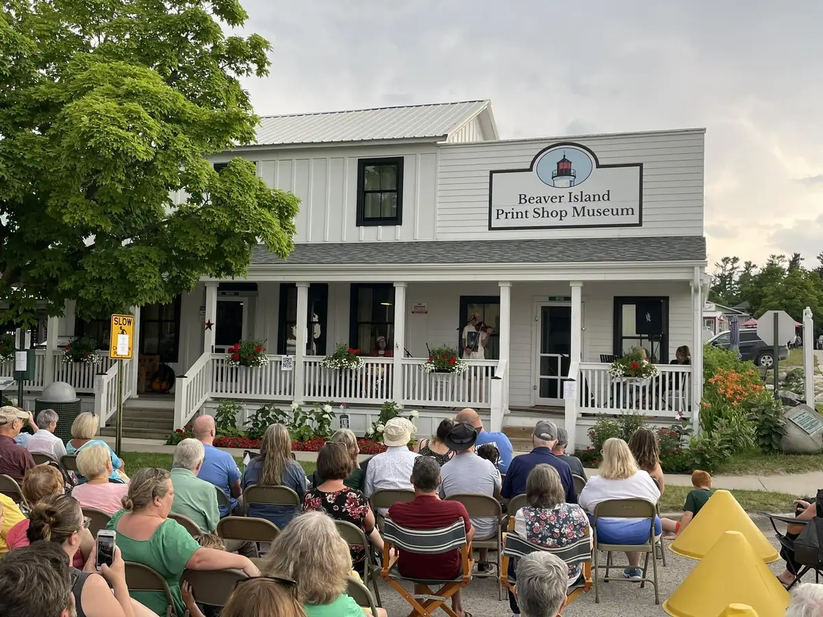 A crowd sits outdoors facing a white building with a porch and a sign reading “Beaver Island Print Shop Museum.”