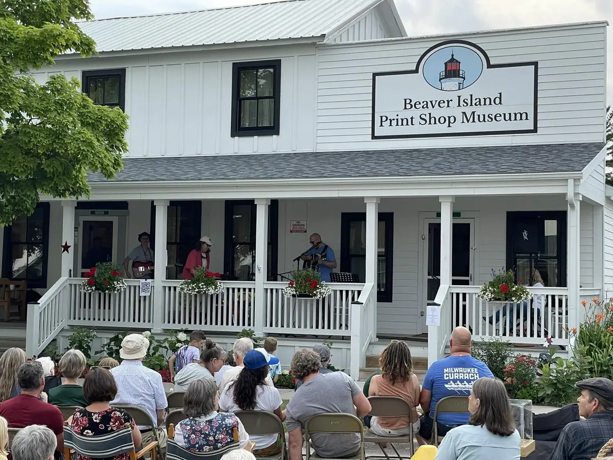 A white building with a museum sign, musicians performing on its porch, and an audience seated outside.