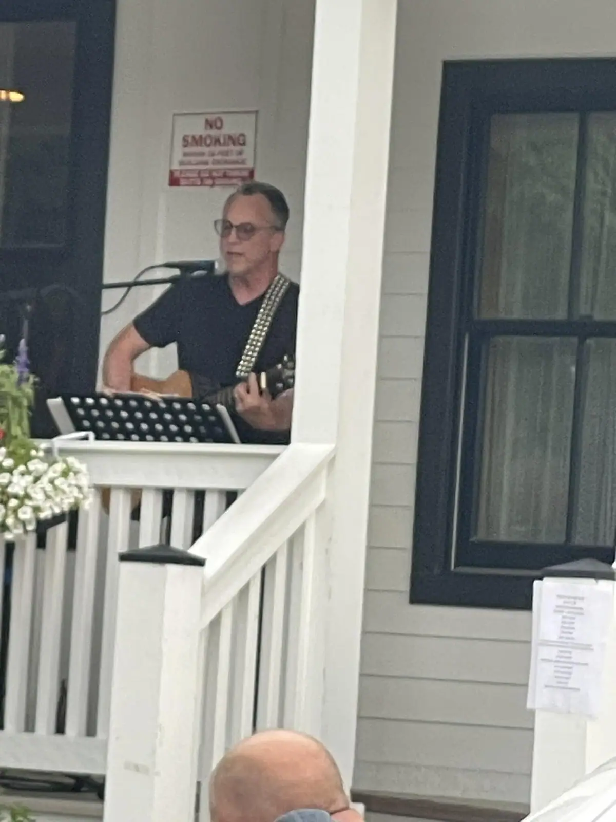 A man plays an acoustic guitar and sings into a microphone on a porch.