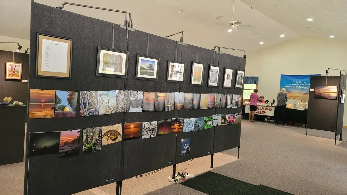 Indoor display of framed and unframed photographs and artwork, with visitors and a jewelry table in the background.