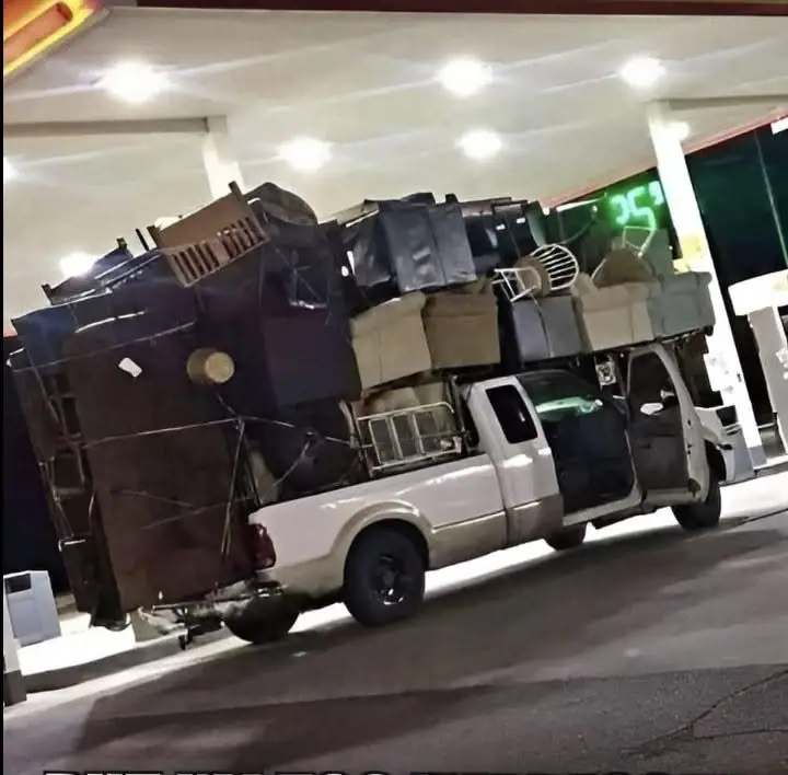 An overloaded white pickup truck carries furniture and household goods beneath a fuel-station canopy.