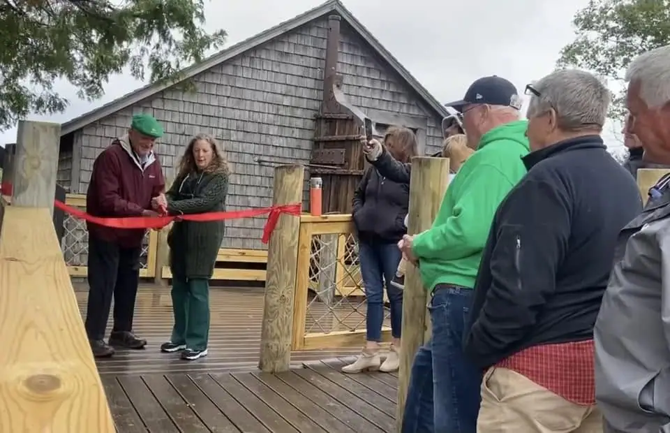 People gather on a wooden deck as two adults cut a red ribbon outside a shingled building.