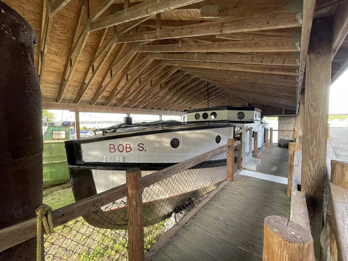 A named boat displayed inside a wooden shed beside a fenced walkway.