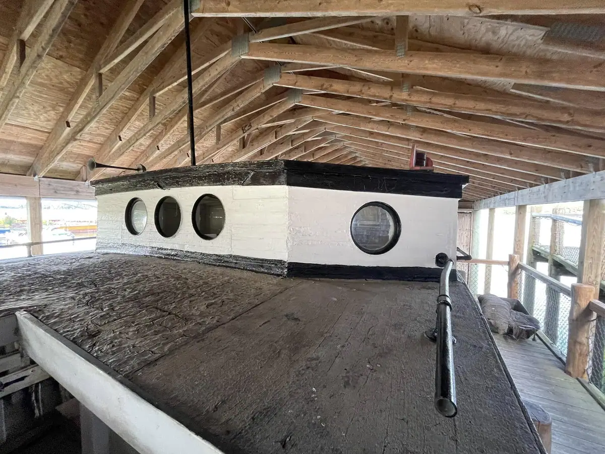 White cabin with round windows and black trim on a wooden deck beneath a timber roof.