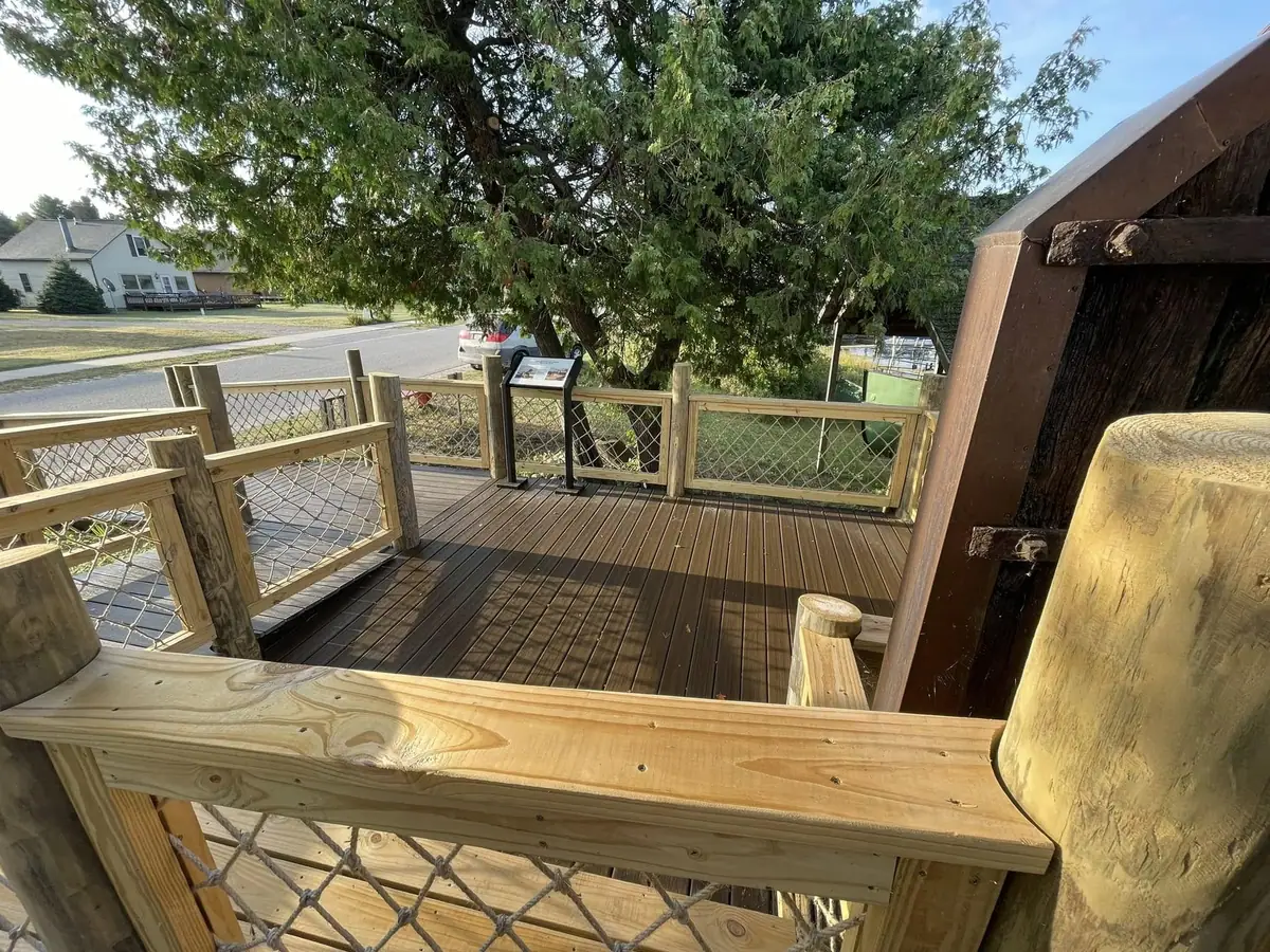A wooden deck with rope-mesh railings overlooks a residential street and houses beneath a large evergreen tree.