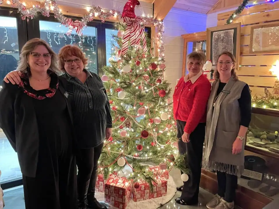 Four women pose beside a decorated Christmas tree and wrapped gifts in an indoor room.