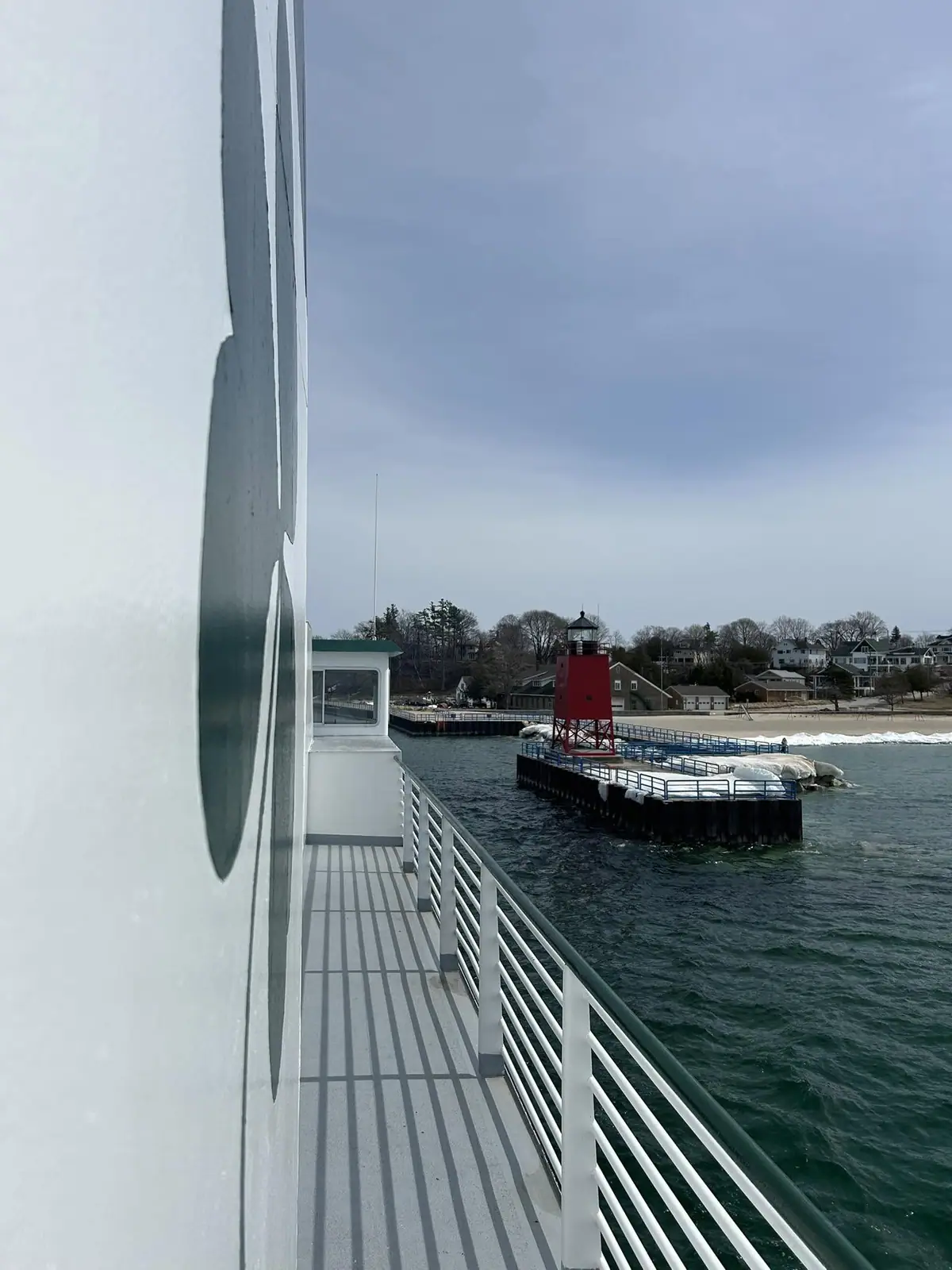 View from a vessel past a red lighthouse on a pier toward a shoreline with houses beneath a cloudy sky.
